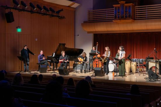A Latin jazz ensemble performs onstage with piano, guitar, bass, saxophone, drums, and percussion as an audience watches.