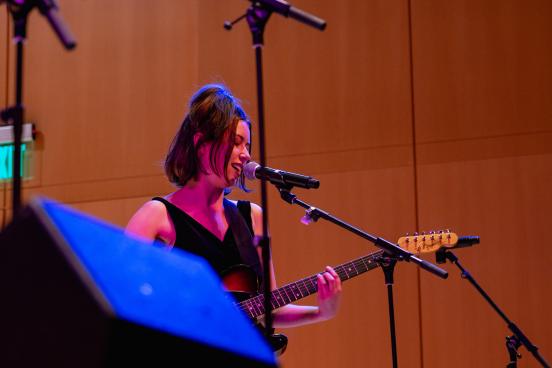 A student musician sings into a microphone while playing an electric guitar during a live indoor performance, illuminated by colorful blue and magenta stage lighting.