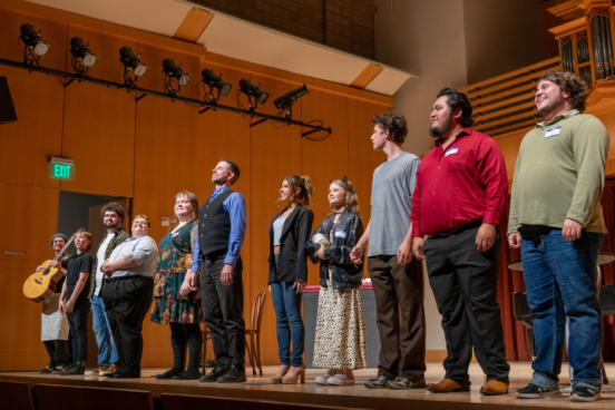 A group of performers stands in a line onstage in a wood-paneled concert hall, facing the audience after a performance.