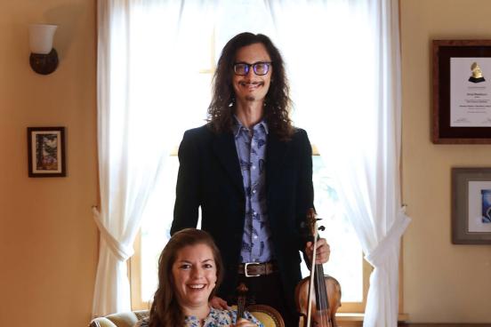 Aaron Westman holding violin standing behind Anna Washburn sitting and holding violin with their dog