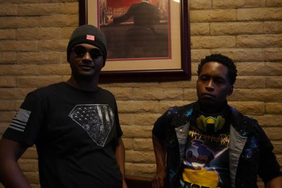 Two people standing indoors against a brick wall, one wearing sunglasses and a beanie, the other wearing a colorful graphic shirt with headphones around the neck.
