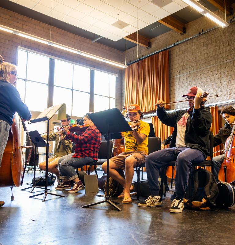 Music students rehearse on violins, cello, and double bass during an ensemble class in a rehearsal room.