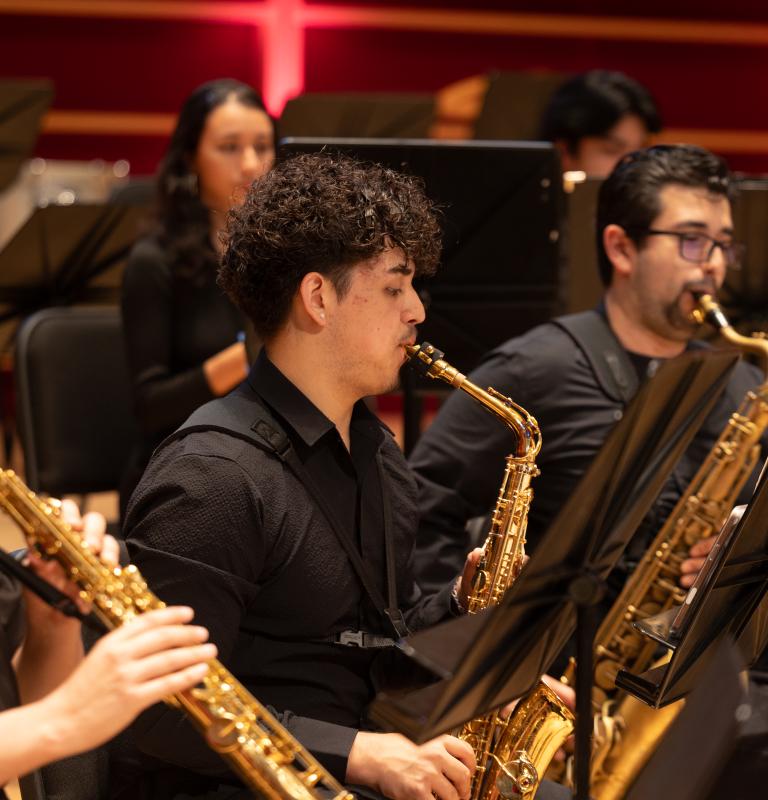 Three saxophonists perform with a wind ensemble, reading sheet music as they play onstage.