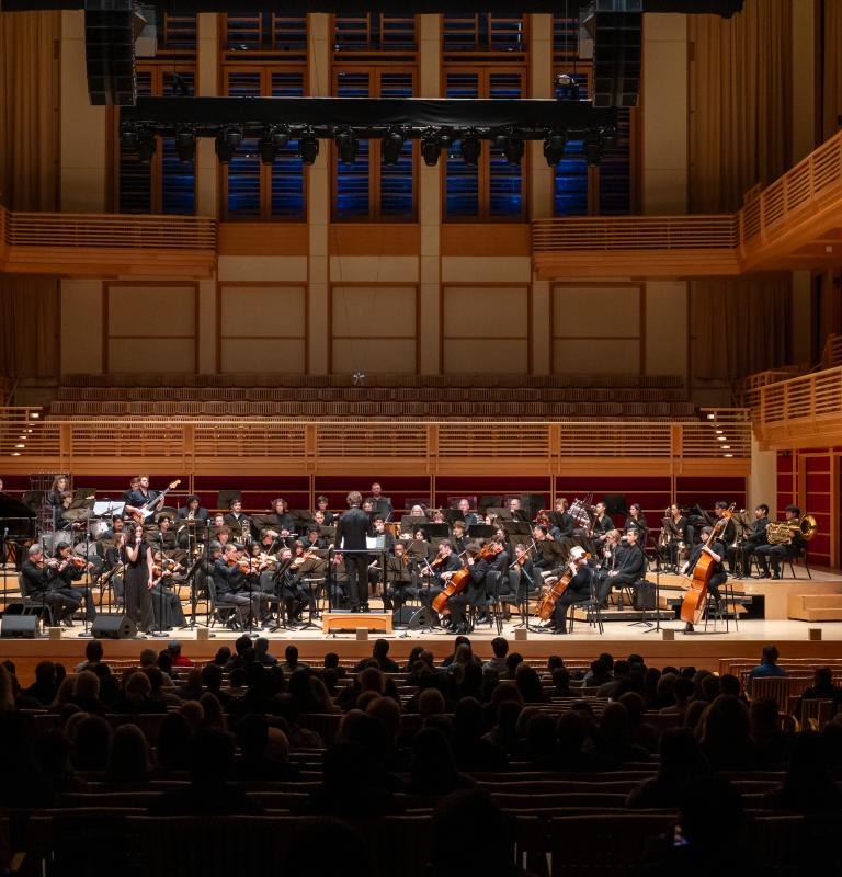 A large orchestra performs onstage in a wood-paneled concert hall as an audience watches from the seats.