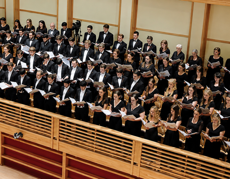 A large choir dressed in black performs from tiered seating in a wood-paneled concert hall while reading from open scores.