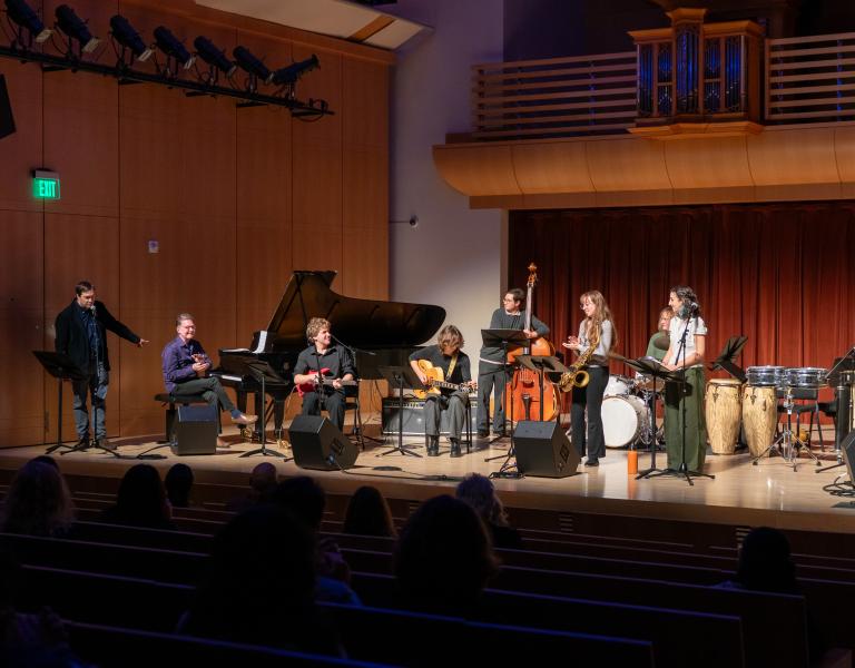 A Latin jazz ensemble performs onstage with piano, guitar, bass, saxophone, drums, and percussion as an audience watches.