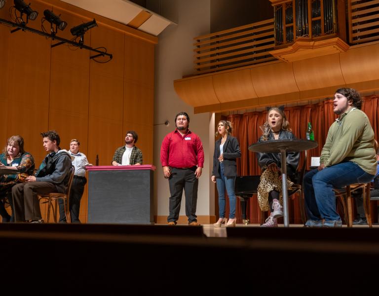 Musical theater performers sing and act onstage in a concert hall, with a bar and café tables forming part of the set.