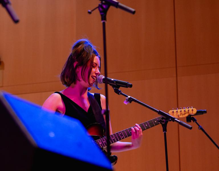 A student musician sings into a microphone while playing an electric guitar during a live indoor performance, illuminated by colorful blue and magenta stage lighting.