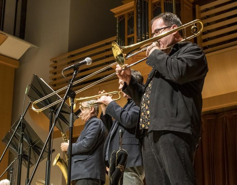 Three musicians playing trombones on stage in a concert hall, with microphones and music stands in front of them