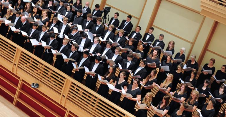 Large choir dressed in formal black attire performing in a concert hall, holding sheet music while singing