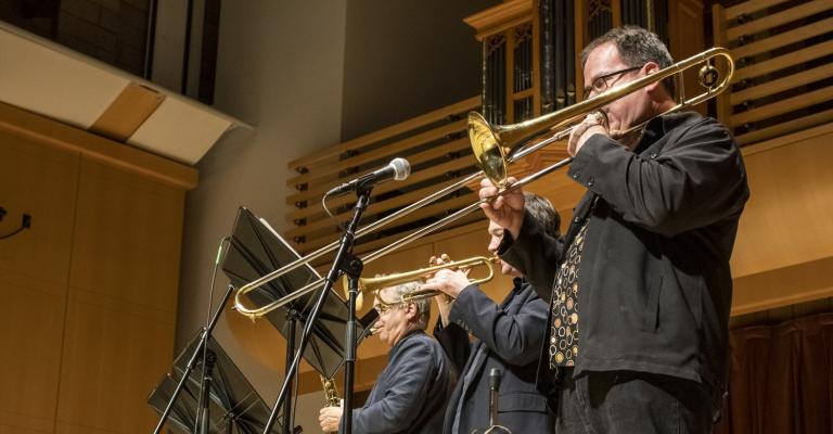 Three musicians playing trombones on stage in a concert hall, with microphones and music stands in front of them