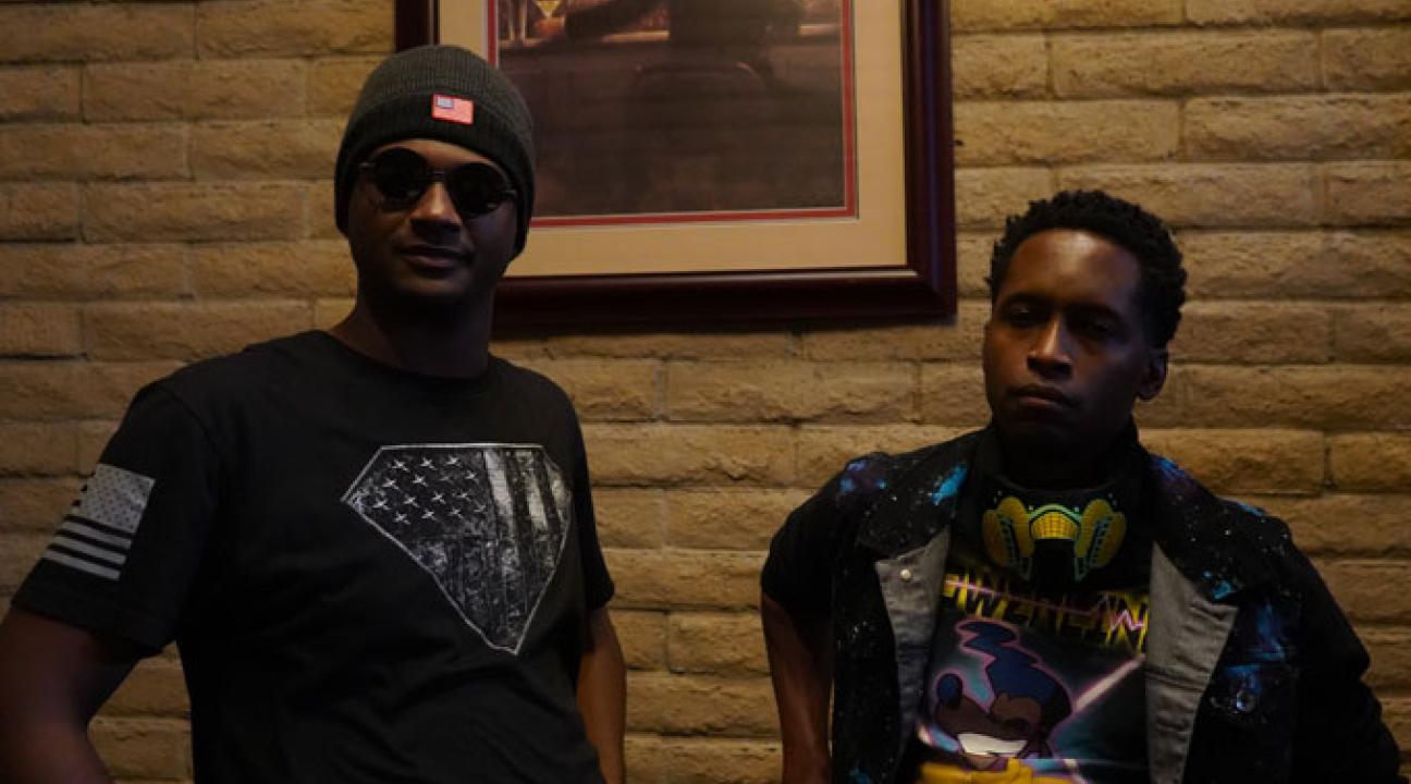 Two people standing indoors against a brick wall, one wearing sunglasses and a beanie, the other wearing a colorful graphic shirt with headphones around the neck.