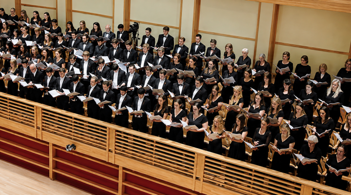 A large choir dressed in black performs from tiered seating in a wood-paneled concert hall while reading from open scores.