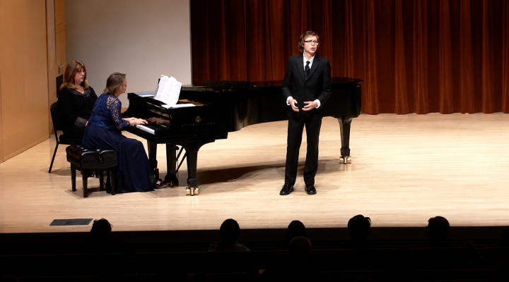 A vocalist in a black suit performs onstage beside a pianist at a grand piano as an audience watches.
