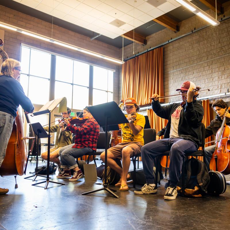 Music students rehearse on violins, cello, and double bass during an ensemble class in a rehearsal room.