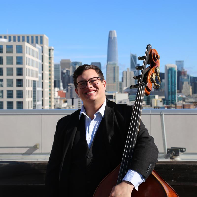 Kody Thiesse smiling in a black suit and white shirt stands on a rooftop holding an upright bass, with the San Francisco skyline and Salesforce Tower visible in the background under a clear blue sky.