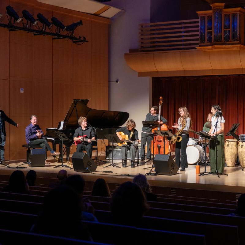 A Latin jazz ensemble performs onstage with piano, guitar, bass, saxophone, drums, and percussion as an audience watches.