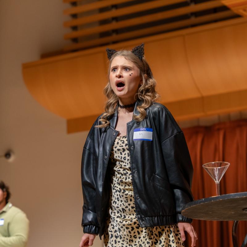 A musical theater performer sings onstage in a leopard-print dress, black jacket, and cat ears during a staged production.