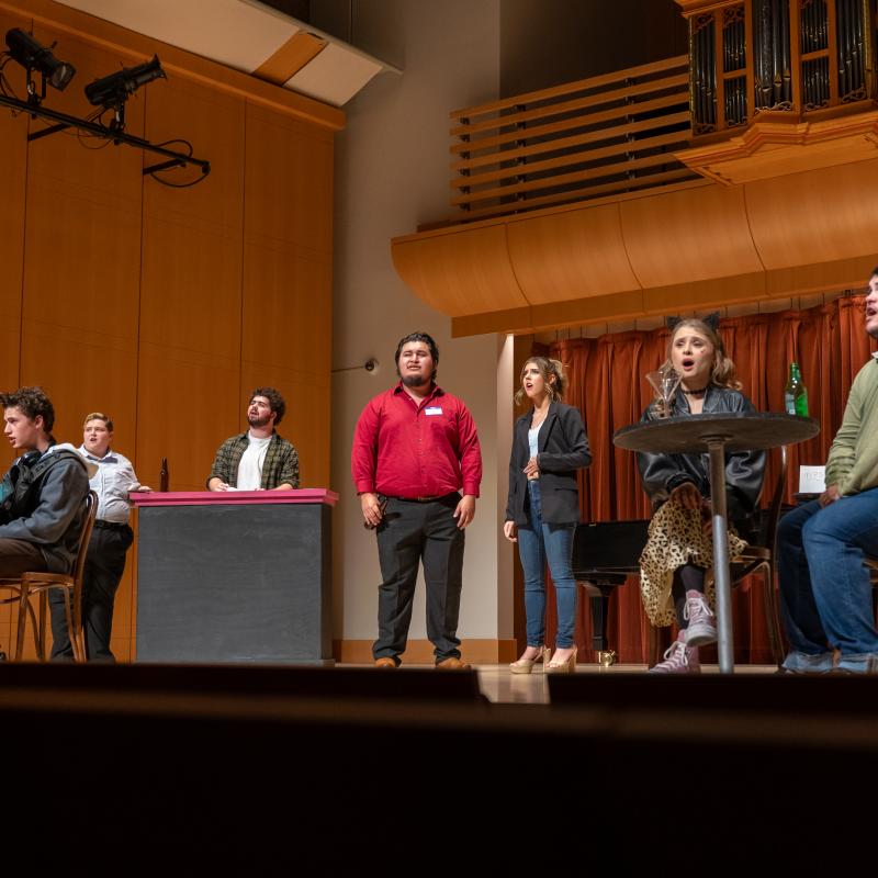 Musical theater performers sing and act onstage in a concert hall, with a bar and café tables forming part of the set.