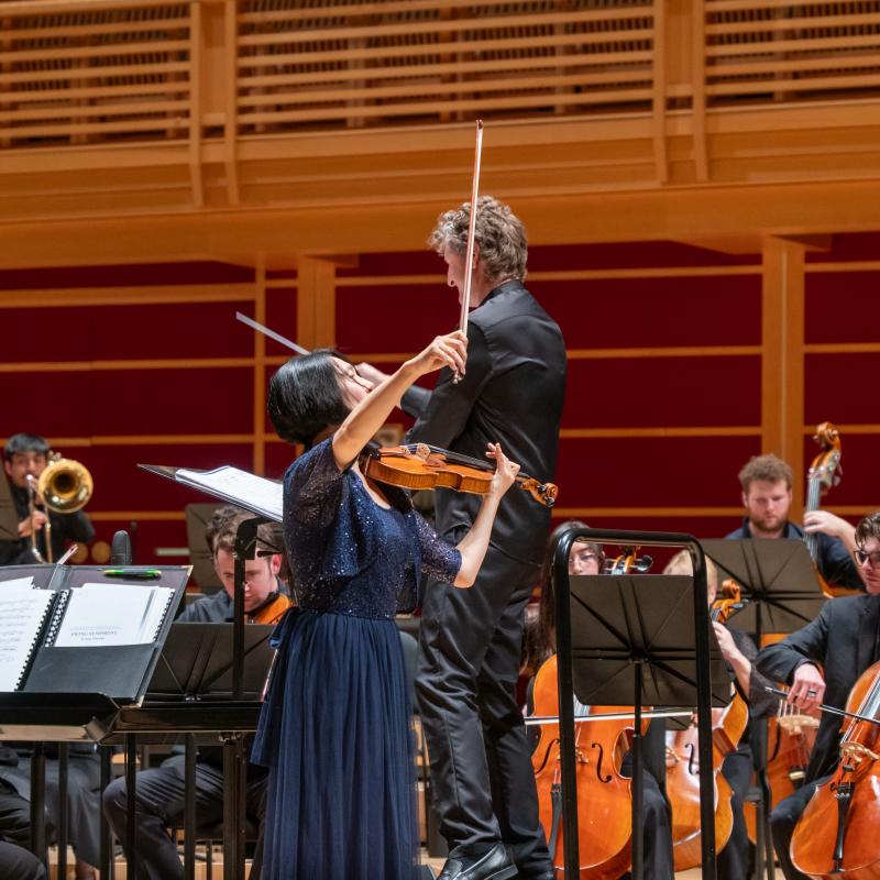 A violin soloist performs alongside a conductor and orchestra in a concert hall.