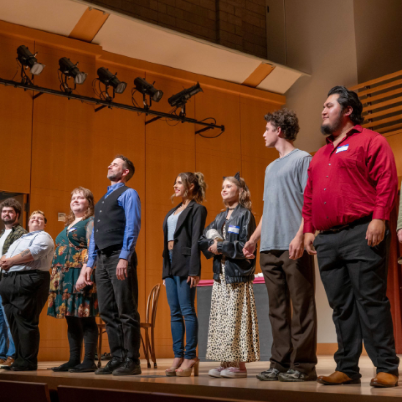 A group of performers stands in a line onstage in a wood-paneled concert hall, facing the audience after a performance.