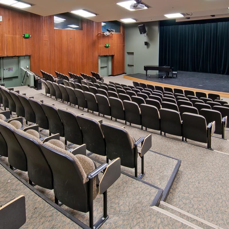 An empty auditorium with tiered seating faces a stage with a grand piano, podium, and closed curtains.