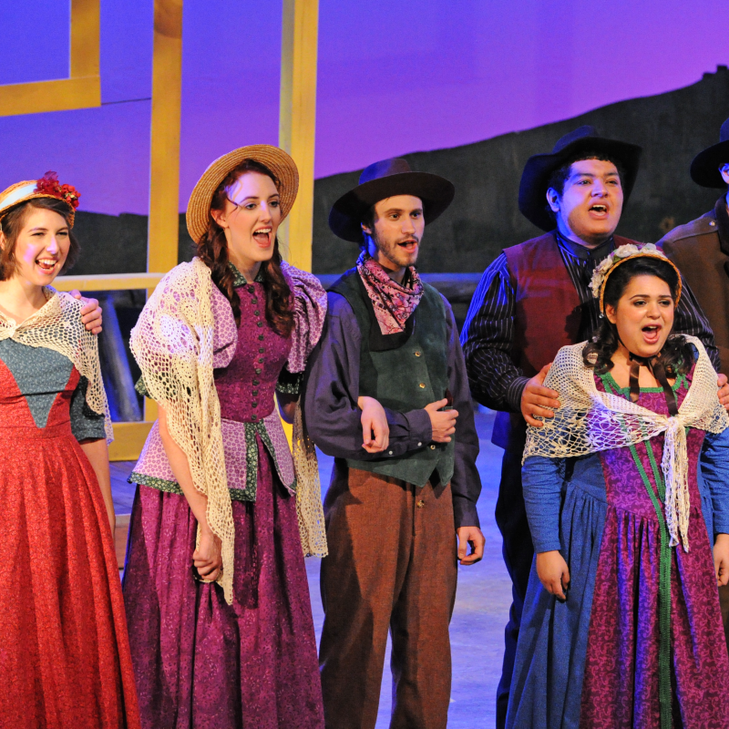 Eight musical theater performers in period costumes sing together onstage during a Western-themed production.
