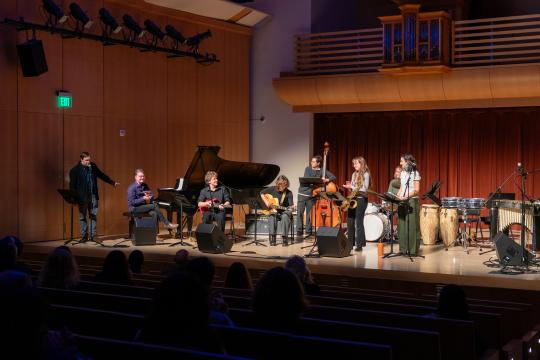 A Latin jazz ensemble performs onstage with piano, guitar, bass, saxophone, drums, and percussion as an audience watches.