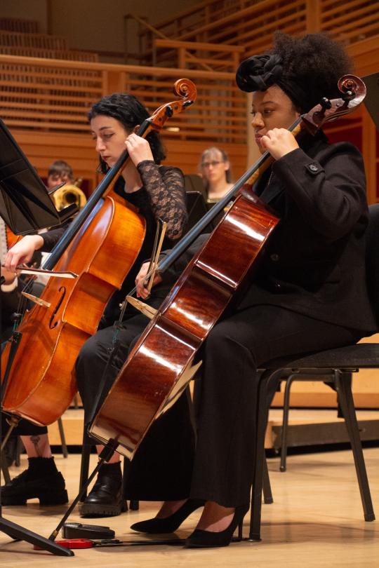 Two musicians playing cellos during an orchestra performance in a concert hall.