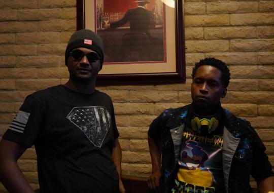 Two people standing indoors against a brick wall, one wearing sunglasses and a beanie, the other wearing a colorful graphic shirt with headphones around the neck.