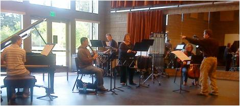 Small ensemble rehearsing indoors with piano, wind instruments, percussion, and a conductor guiding the musicians