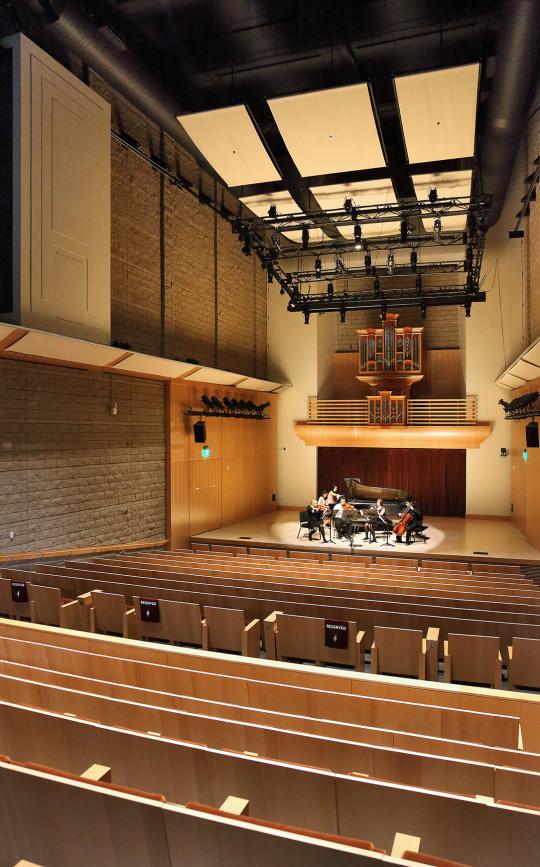 Small musical ensemble performing on stage with a grand piano inside a modern concert hall featuring wooden seating, high ceilings, and an organ in the back