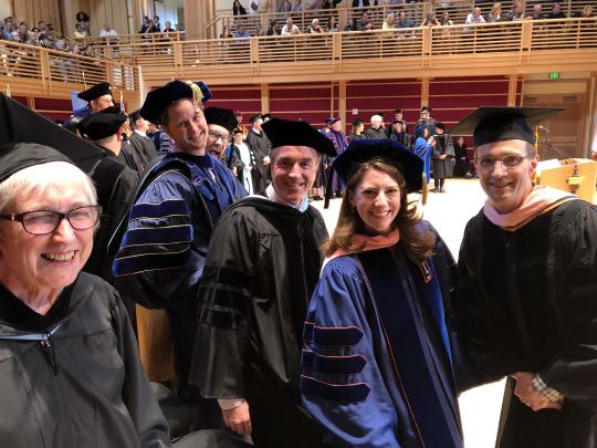 Group of faculty members in academic regalia smiling during a commencement ceremony inside an auditorium filled with people