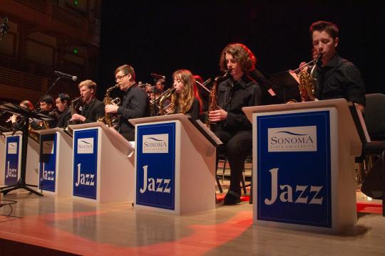 Sonoma State University Jazz Ensemble performing on stage, with musicians playing saxophones and brass instruments behind stands labeled ‘Sonoma State University Jazz