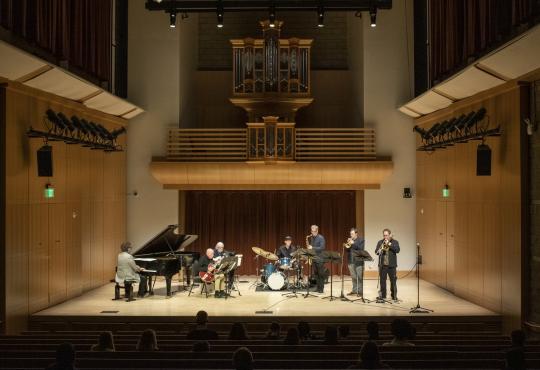 Small jazz ensemble performing on stage in a concert hall, featuring piano, guitar, drums, and brass instruments with an organ in the background