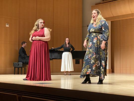 Two performers singing on stage, one in a red dress and the other in a floral dress, accompanied by a pianist and another person standing near the piano.