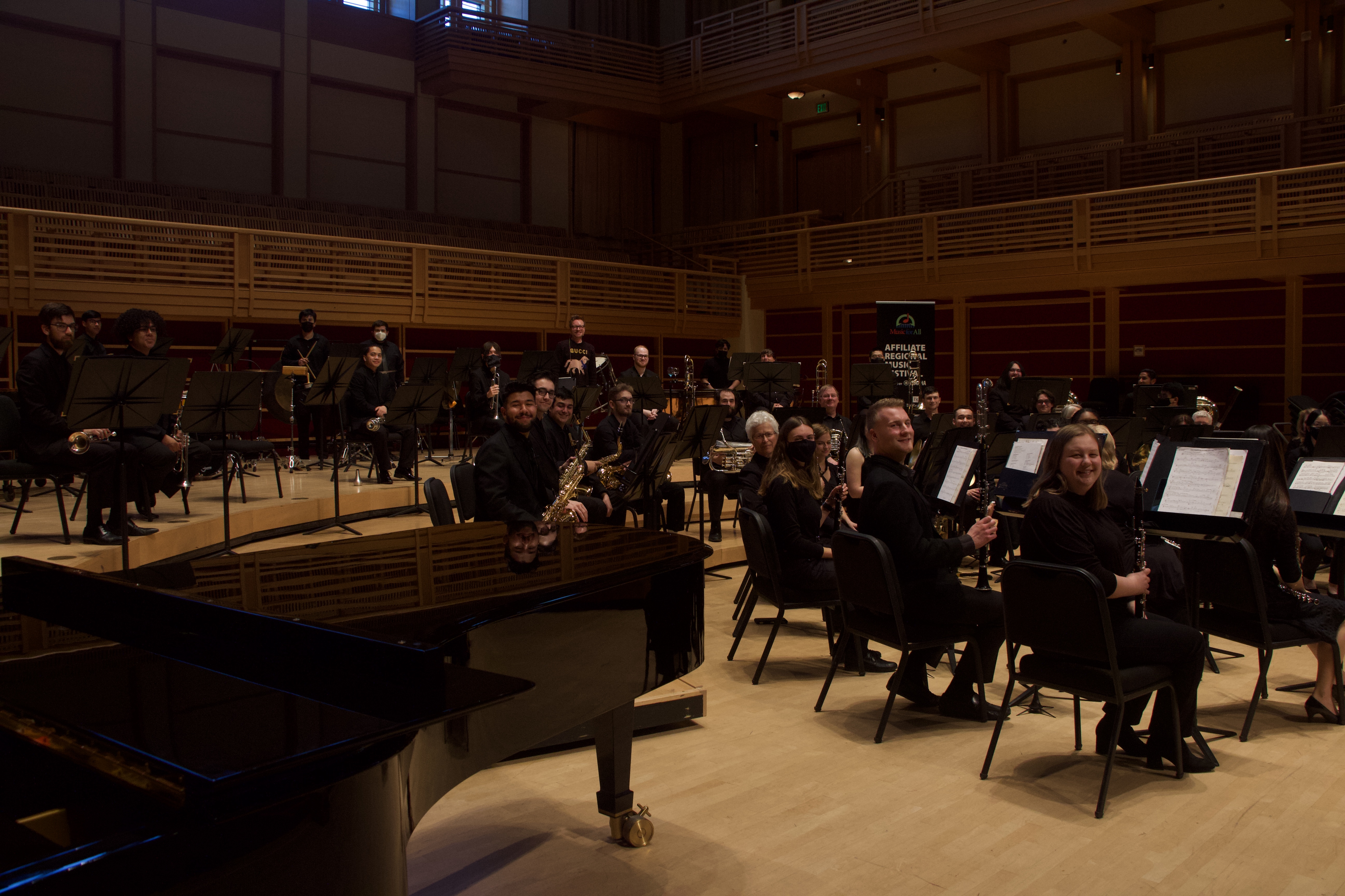 The Symphonic Wind Ensemble seated, posing and smiling on stage with John Mackey