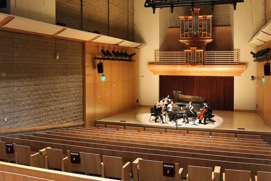 Small musical ensemble performing on stage with a grand piano inside a modern concert hall featuring wooden seating, high ceilings, and an organ in the back
