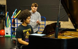 Jomei Greer practicing at piano with instructor behind