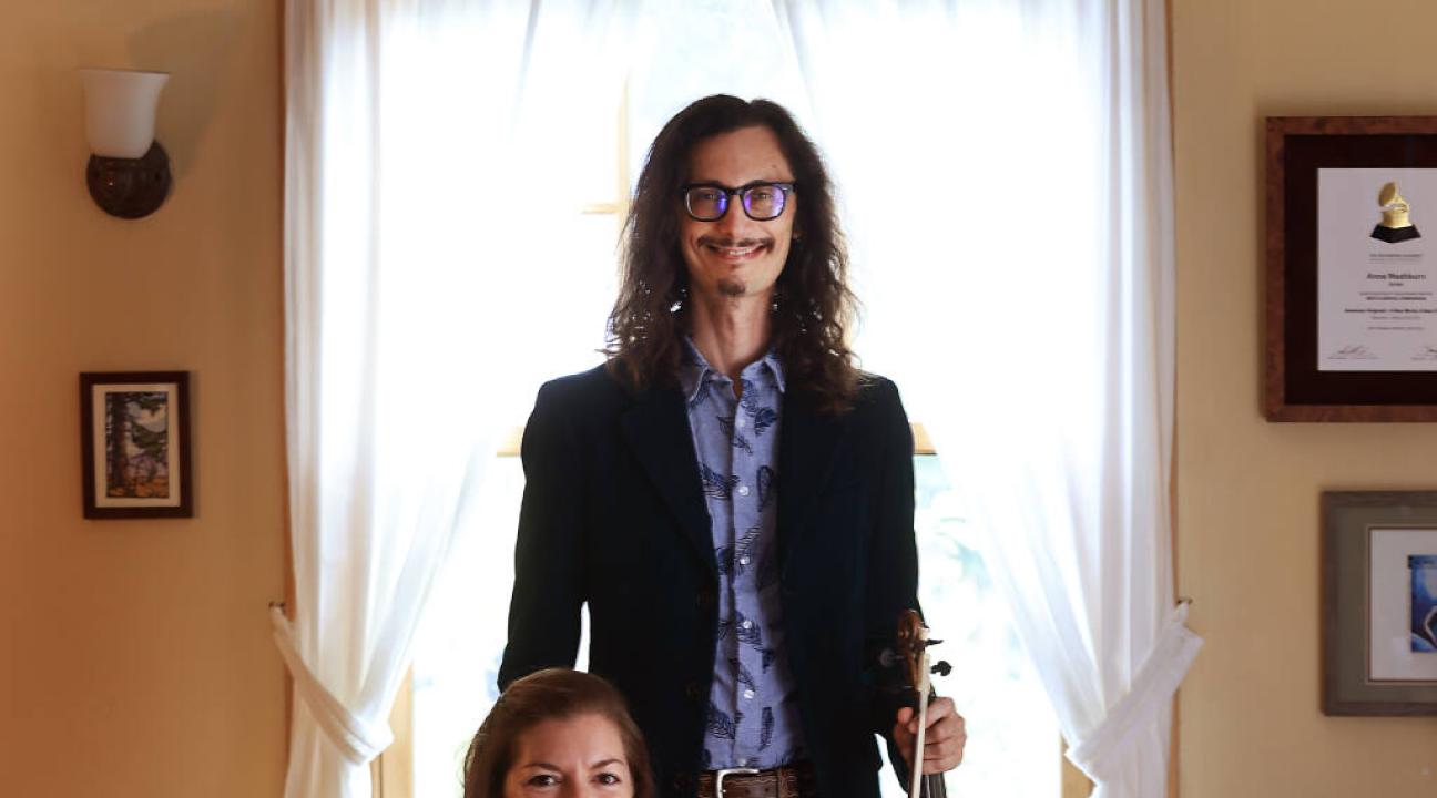 Aaron Westman holding violin standing behind Anna Washburn sitting and holding violin with their dog