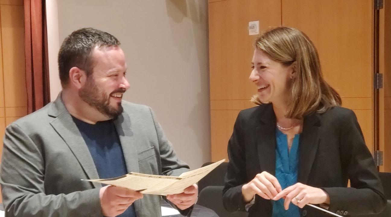 Justin Montigne holding paper and Jenny Bent holding a baton looking at each other and laughing
