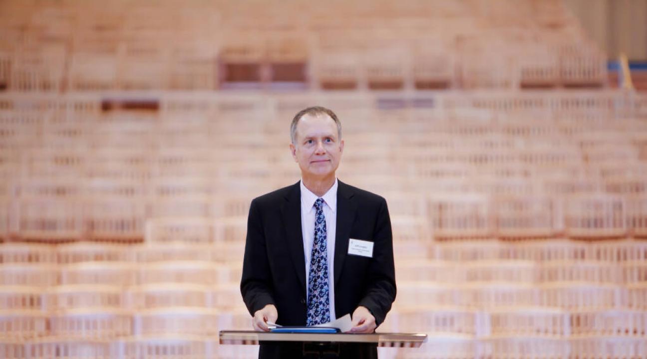 Jeff Langley dressed in a suit and standing at a podium with empty wooden seats behind him