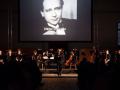 conductor standing in front of ensemble in front of slide show with picture of Viktor Ulmann