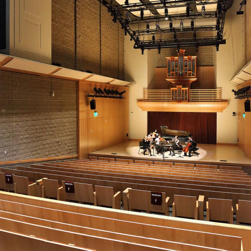 Small musical ensemble performing on stage with a grand piano inside a modern concert hall featuring wooden seating, high ceilings, and an organ in the back