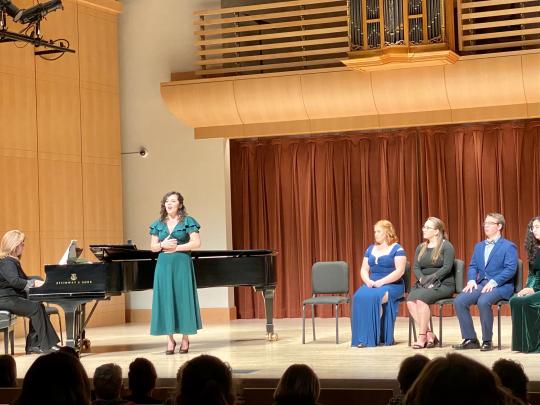 Four students sitting on stage while one student sings in front of a piano and pianist with the silhouette of the audience in the foreground