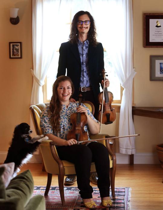 Aaron Westman holding violin standing behind Anna Washburn sitting and holding violin with their dog