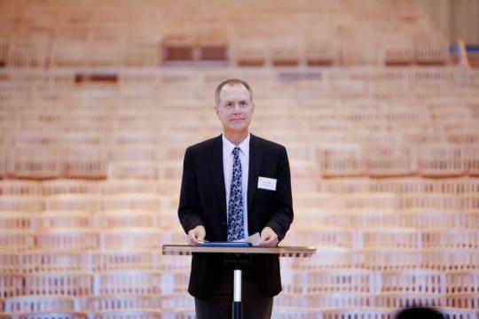 Jeff Langley dressed in a suit and standing at a podium with empty wooden seats behind him