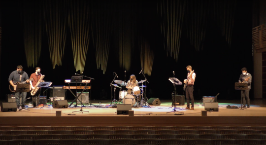 Band playing on stage at Weill Hall with drums, guitars, saxophone, and lighting in the background