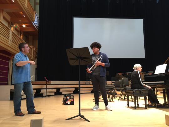 Instructor teaching trumpet student on stage while piano accompanist sits to the right