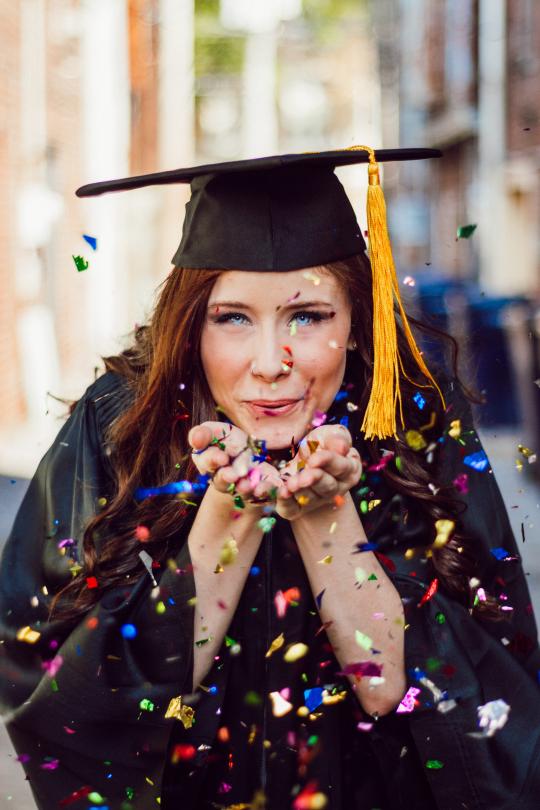 a girl in graduation garb and mortarboard blowing colorful confetti